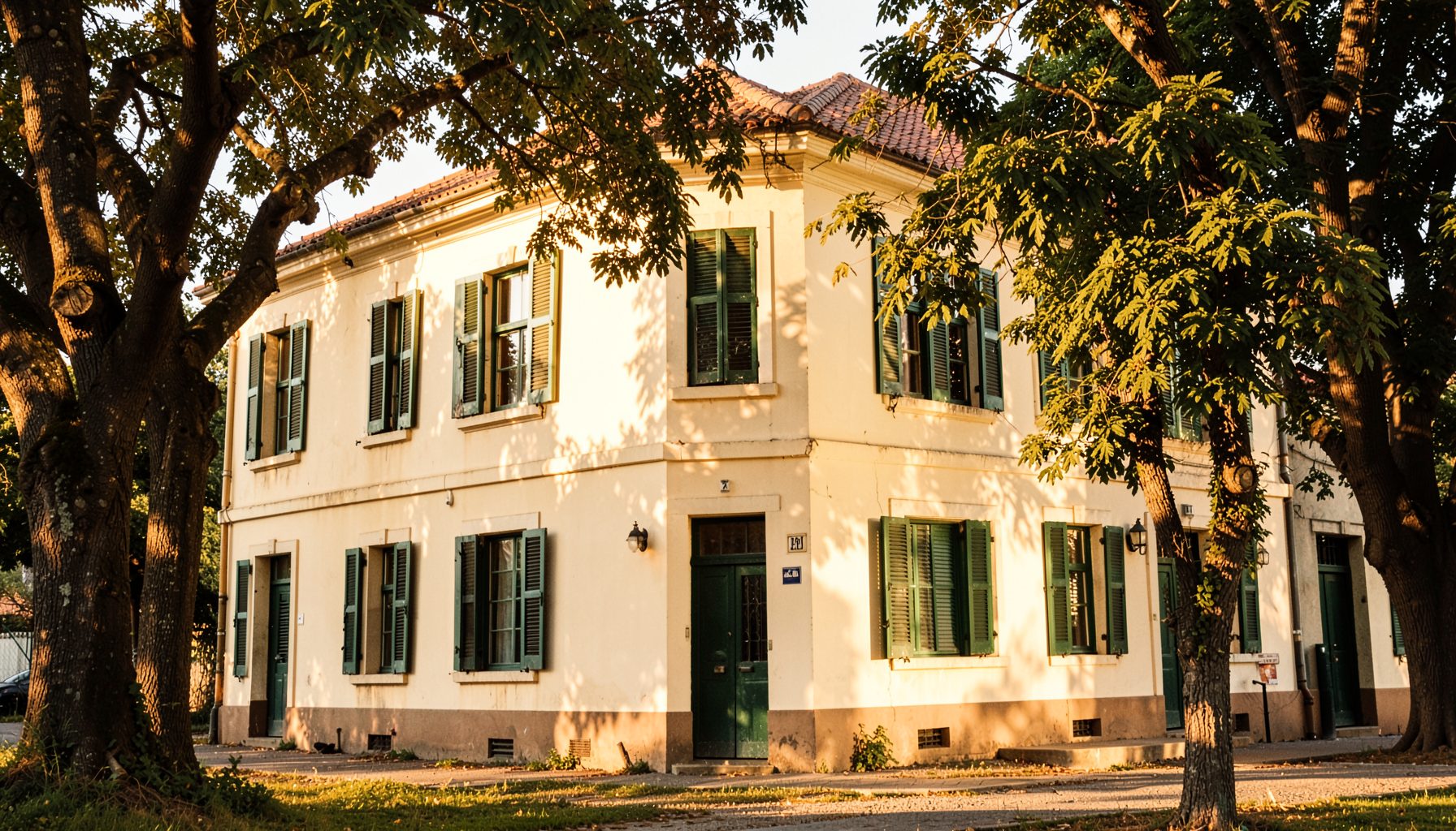 A warm, sunlit view of a traditional school building with cream-colored walls and green window shutters, framed by mature trees, conveying nostalgia and community pride.